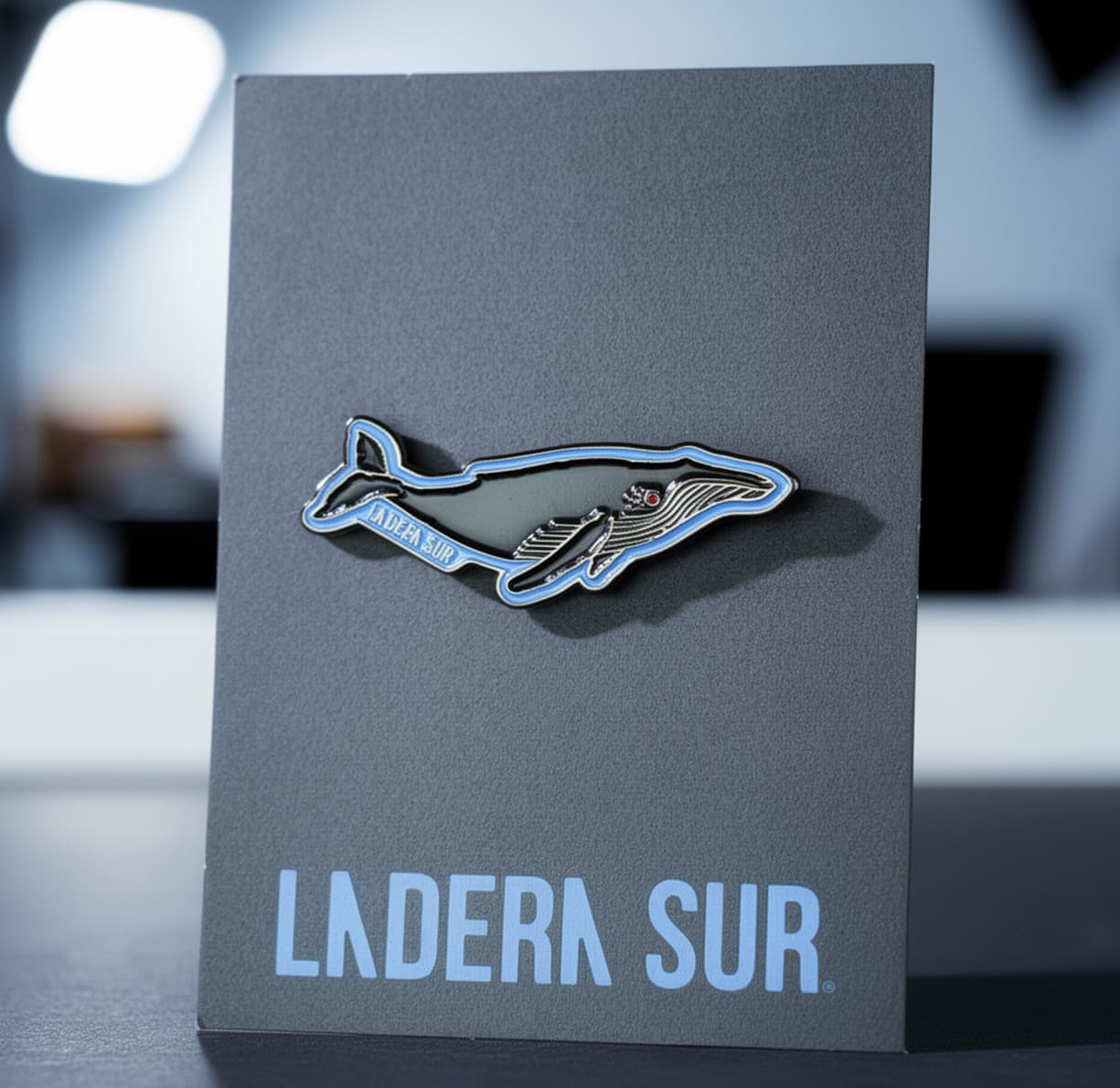 Pin metálico esmaltado con forma de Ballena Jorobada. Cuerpo gris oscuro con detalles en azul claro y ojo rojo. Montado sobre tarjeta gris con logo Ladera Sur en letras celestes.